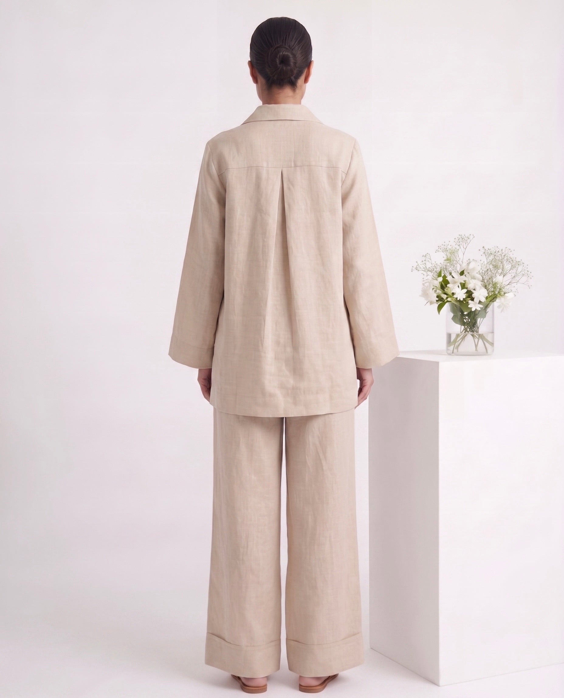 A model viewed from the back wearing a light beige linen two-piece set consisting of a long-sleeve button-up jacket with a central pleat and matching wide-leg trousers. The model stands on a white pedestal next to a small vase of white flowers against a clean white background.