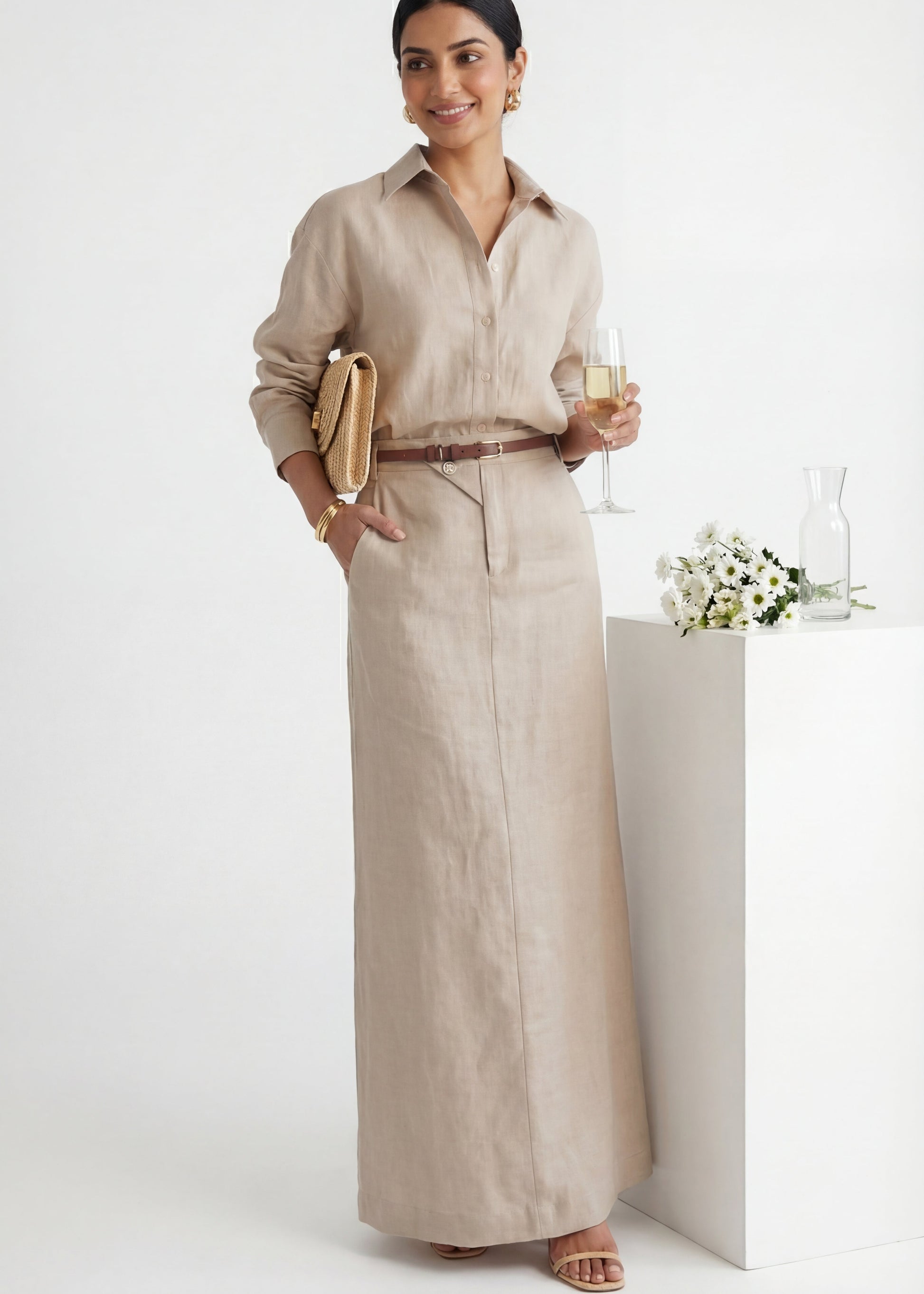 Woman in a beige outfit holding a glass of wine and a clutch, standing against a white background.