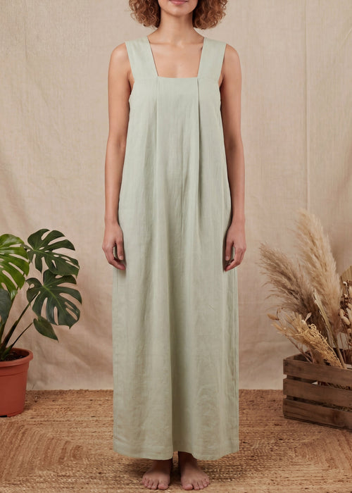 Woman wearing a light green dress standing in front of a beige curtain with plants on either side.