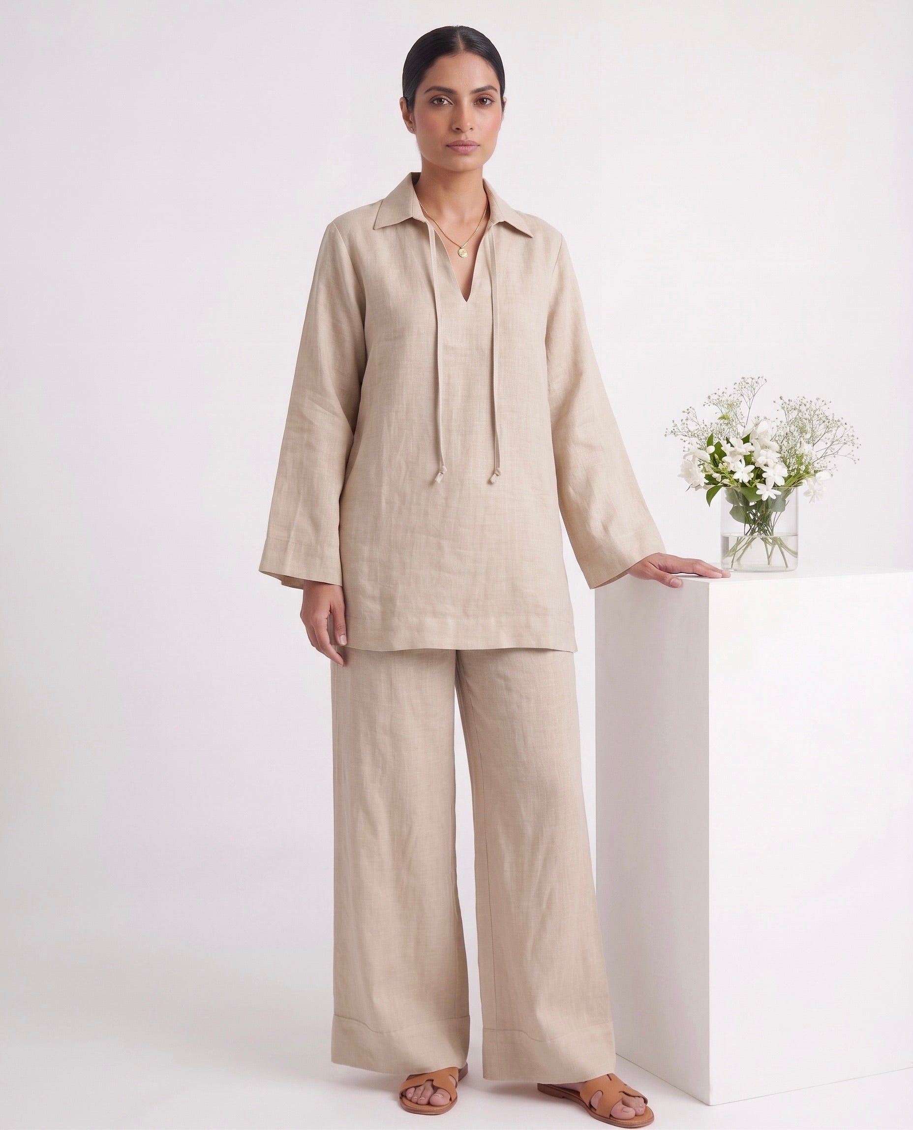 A woman modeling a light beige linen two-piece set consisting of a long-sleeve button-up top with drawstrings and matching wide-leg trousers, standing in a studio next to a white pedestal with a small vase of flowers.