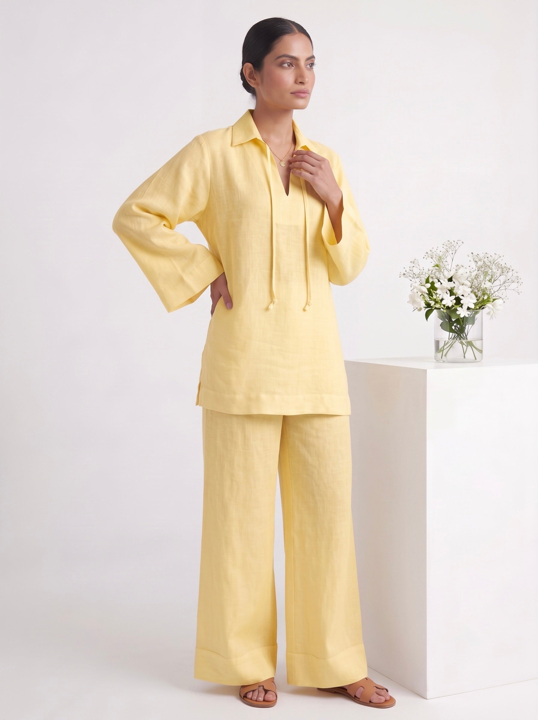 A woman modeling a light yellow two-piece pajama set consisting of a long-sleeve collared top with drawstrings and matching wide-leg pants, standing in a studio next to a white pedestal with a small vase of flowers.