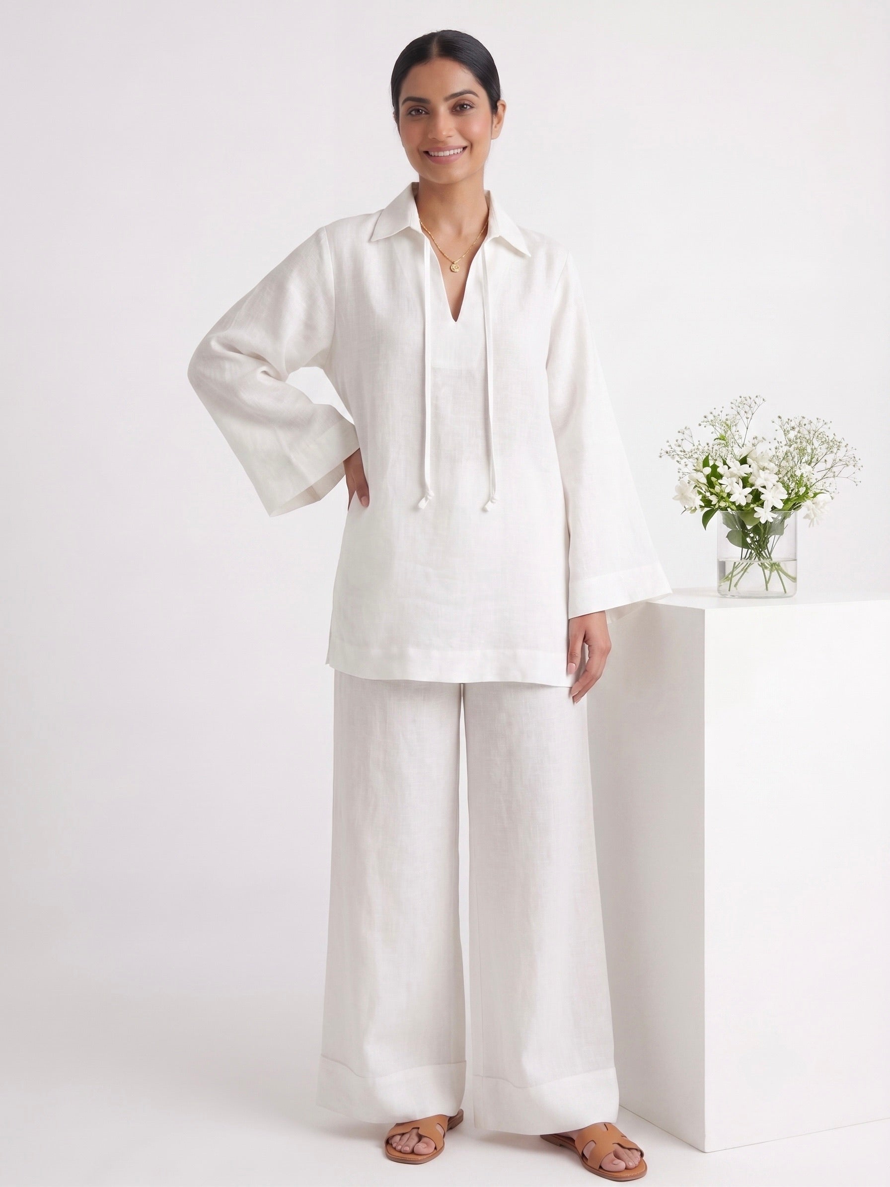 A woman modeling a white two-piece linen set consisting of a long-sleeve collared top with drawstrings and matching wide-leg pants, standing in a bright white studio next to a white pedestal with a small vase of flowers.