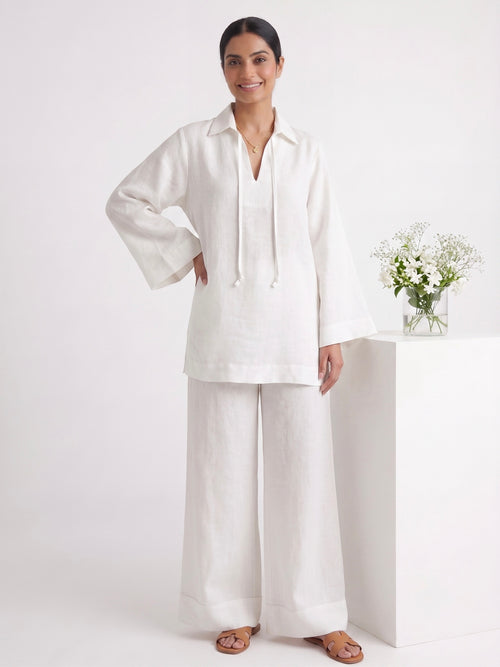 A woman modeling a white two-piece linen set consisting of a long-sleeve collared top with drawstrings and matching wide-leg pants, standing in a bright white studio next to a white pedestal with a small vase of flowers.