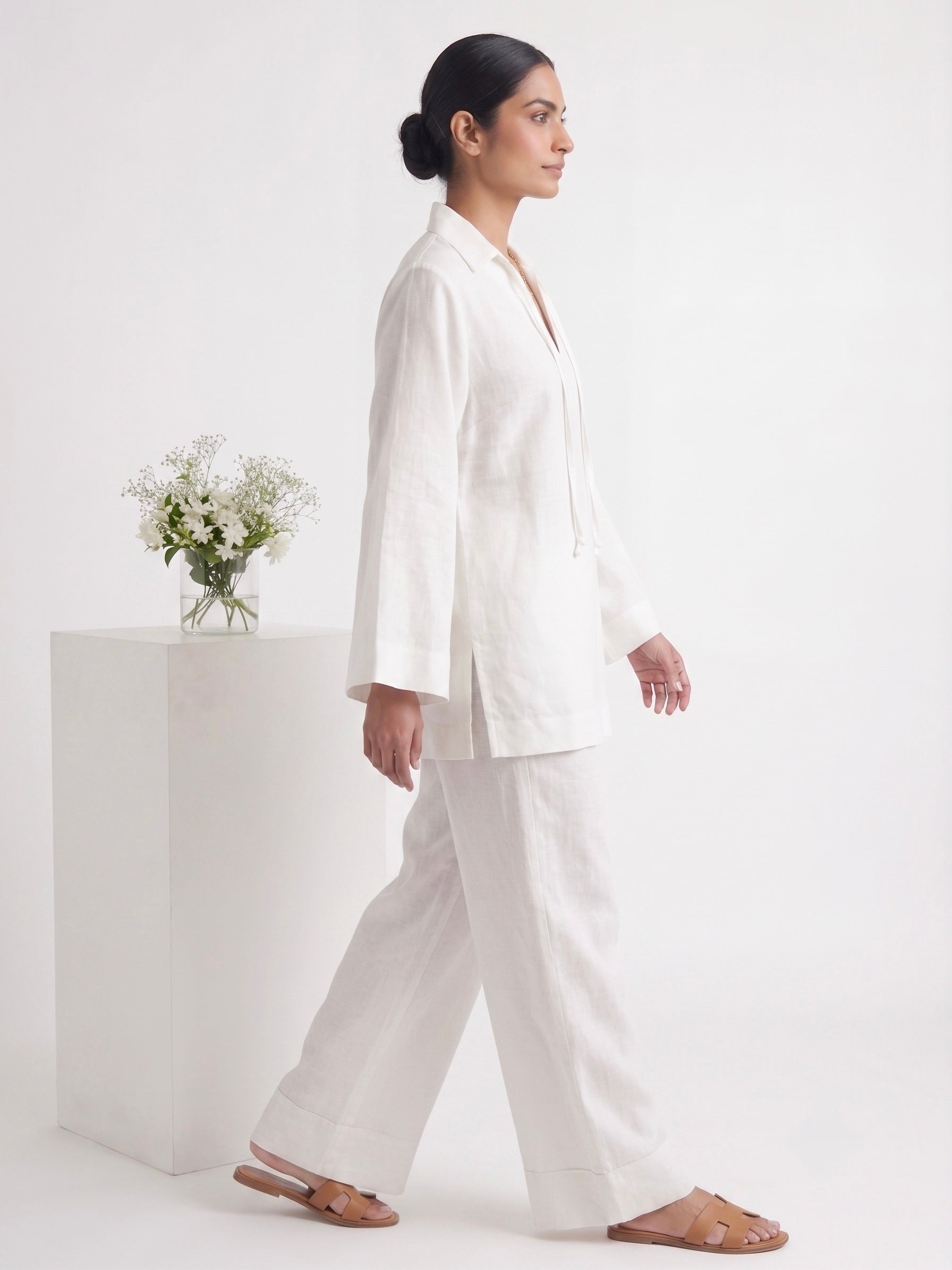 A woman in a white two-piece linen set consisting of a long-sleeve button-up blazer and matching wide-leg trousers, walking in profile. She is wearing brown leather flat sandals and standing in a bright, minimalist studio with a white pedestal holding a small vase of flowers.