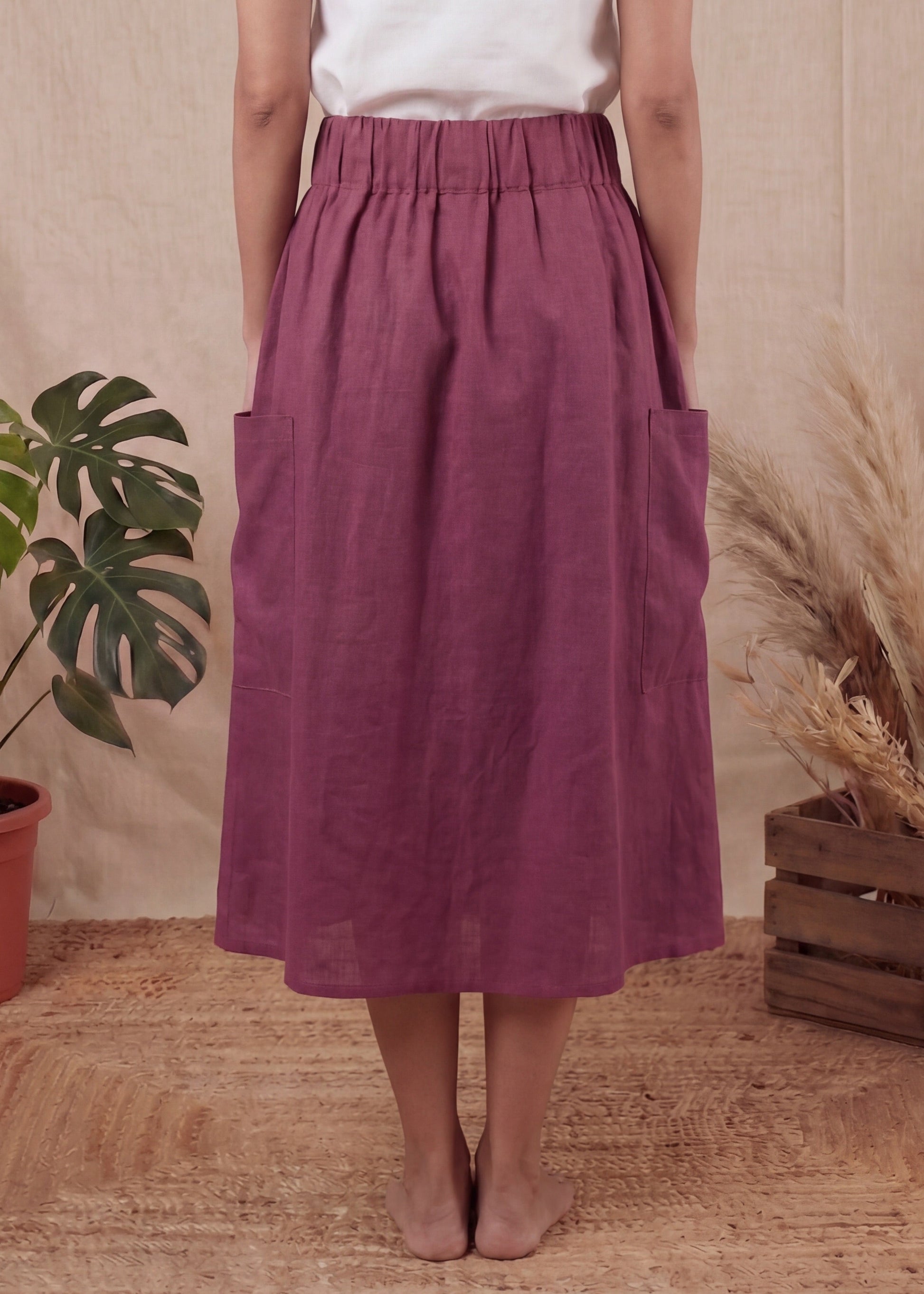 Person wearing a white top and purple skirt standing in front of a beige curtain with plants on either side.