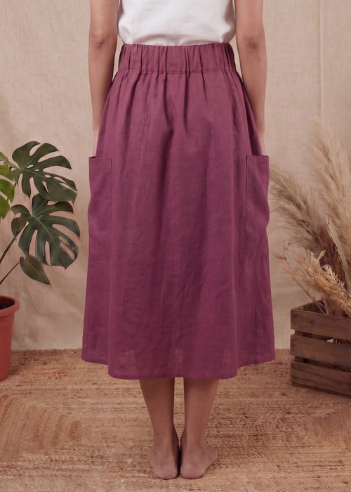 Person wearing a white top and purple skirt standing in front of a beige curtain with plants on either side.