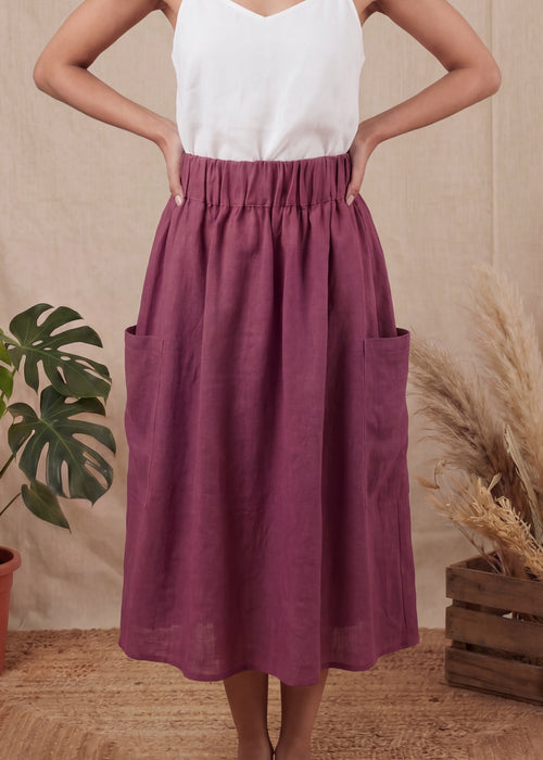 Woman wearing a white top and purple skirt standing in front of a beige curtain with plants on either side.